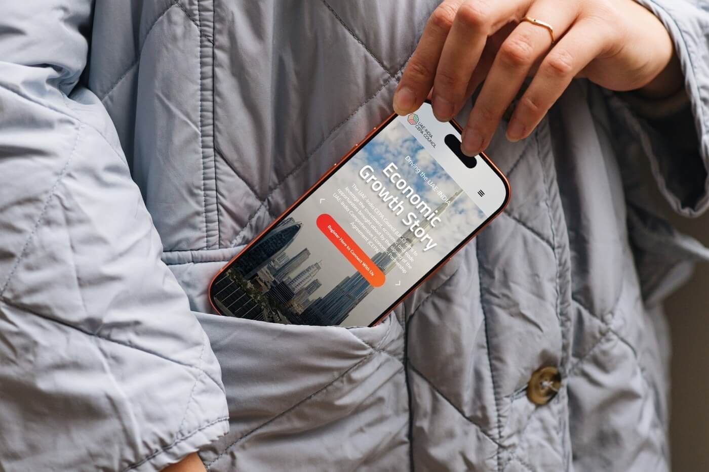 A hand pulling a phone out of a quilted jacket pocket. The screen shows the mobile home page, headed “Economic Growth Story” over a city skyline.