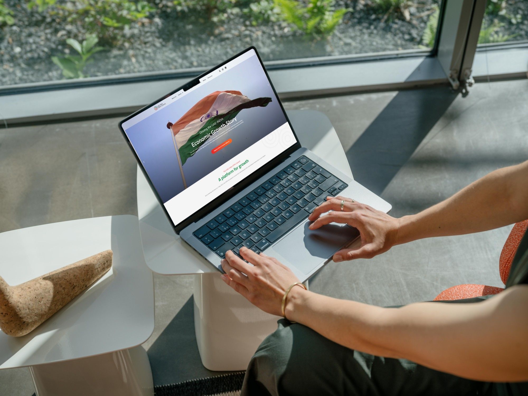 Hands typing on a laptop on a white side table by a window. The screen shows the CEPA Council website’s home page: “Driving the UAE-India Economic Growth Story” over a waving Indian flag.